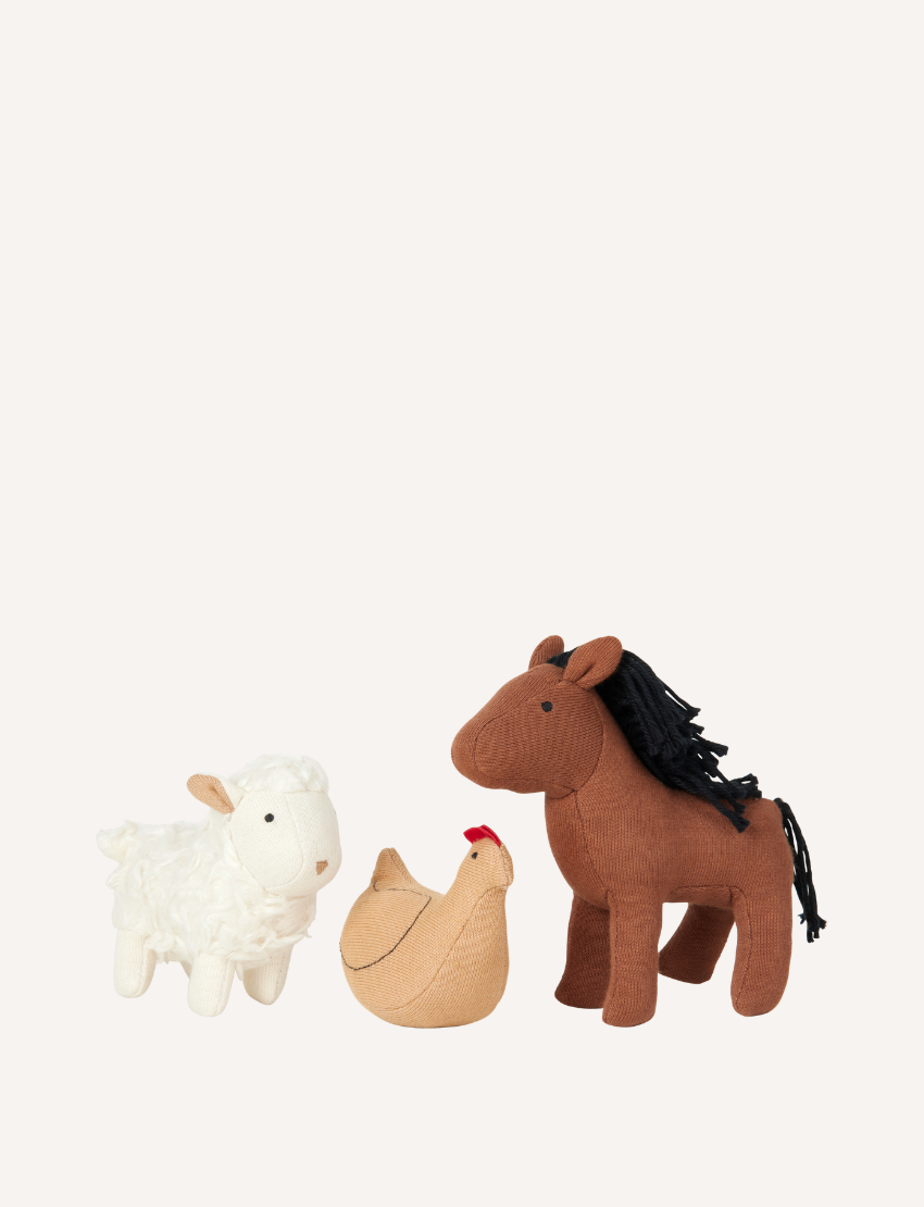 On a quilted surface sits the Pocket Friend - Farm Animals from Fabelab: a plush barn toy with an open door. Surrounding the barn are small plush figures, including a horse, chicken, child, and sheep. The interior of the barn is adorned with farm-themed designs and patterns.