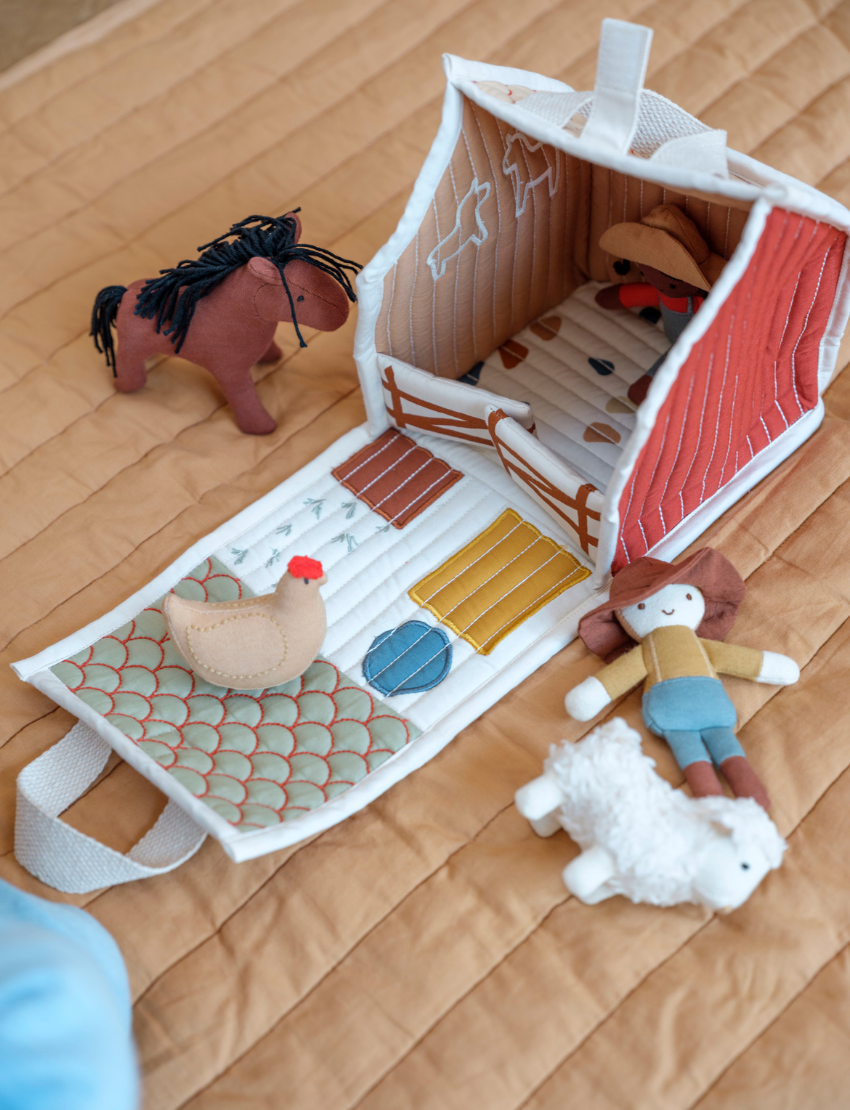 On a quilted surface sits the Pocket Friend - Farm Animals from Fabelab: a plush barn toy with an open door. Surrounding the barn are small plush figures, including a horse, chicken, child, and sheep. The interior of the barn is adorned with farm-themed designs and patterns.