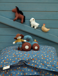 On a quilted surface sits the Pocket Friend - Farm Animals from Fabelab: a plush barn toy with an open door. Surrounding the barn are small plush figures, including a horse, chicken, child, and sheep. The interior of the barn is adorned with farm-themed designs and patterns.