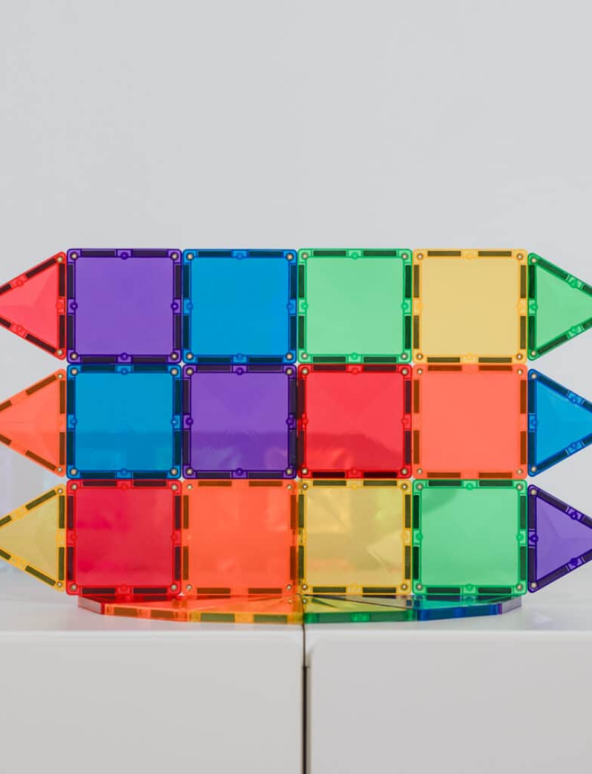 The Connetix Tiles Rainbow Mini Pack 24-Piece set is on display. The packaging features a purple and beige design with illustrations of vibrant magnetic tiles creating a rocket and various shapes. It includes 24 pieces and is suitable for children aged 3 and older.