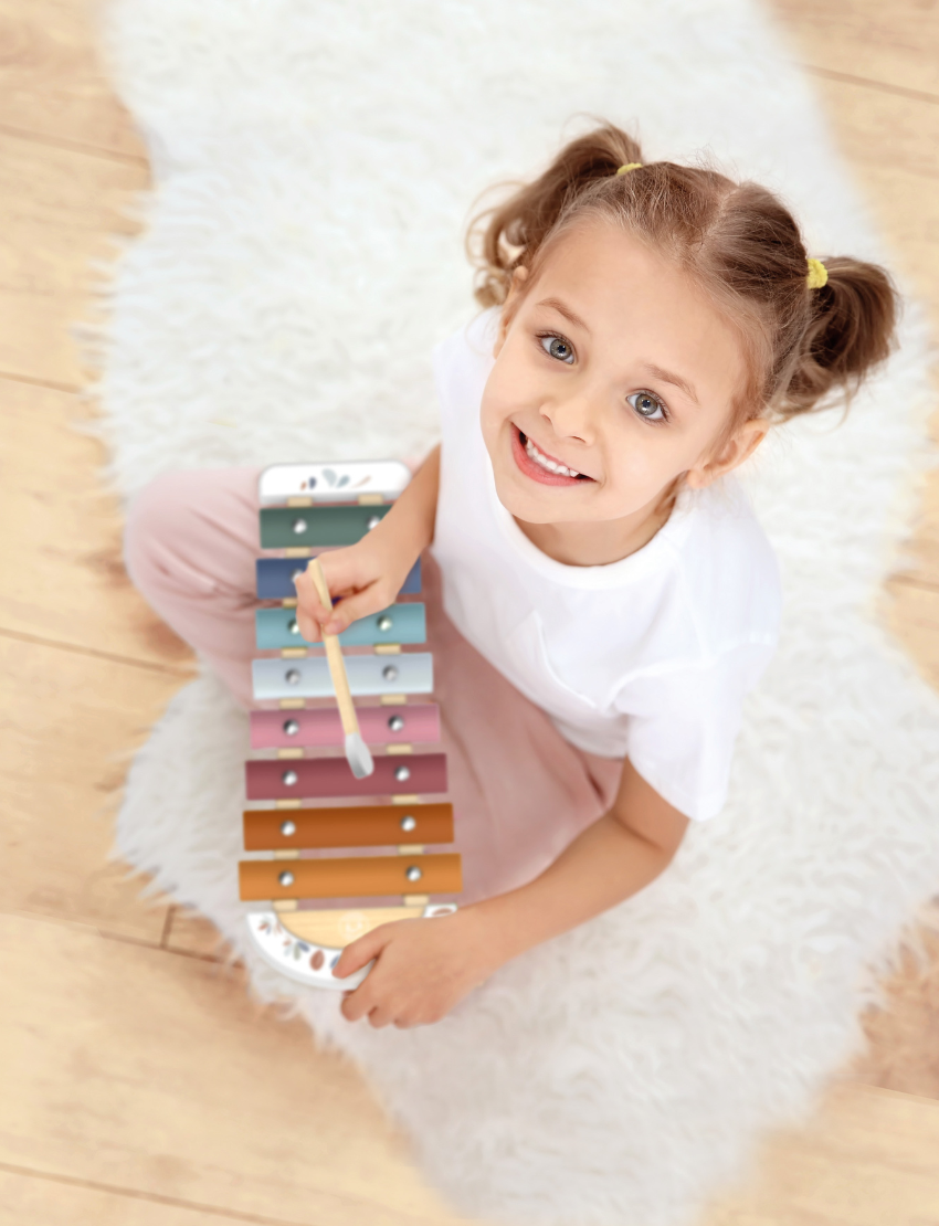 The Speedy Monkey Xylophone, featuring eight vibrant metal bars in shades of orange, yellow, green, blue, and pink, rests on a white surface. In front of the instrument lies a wooden mallet with a rounded white end.