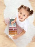 The Speedy Monkey Xylophone, featuring eight vibrant metal bars in shades of orange, yellow, green, blue, and pink, rests on a white surface. In front of the instrument lies a wooden mallet with a rounded white end.