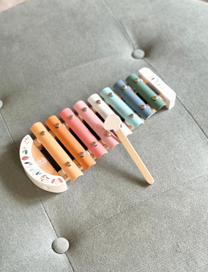 The Speedy Monkey Xylophone, featuring eight vibrant metal bars in shades of orange, yellow, green, blue, and pink, rests on a white surface. In front of the instrument lies a wooden mallet with a rounded white end.