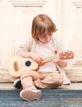 A young child with light brown hair sits on the ground playing a Speedy Monkey Ukulele. The child is dressed in a beige sweater, light pink shirt, and tan pants, with a light-colored wooden door in the background.