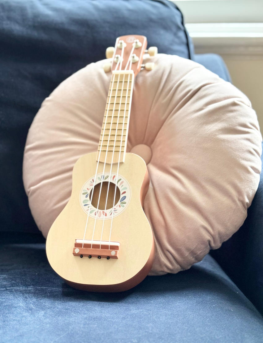 A young child with light brown hair sits on the ground playing a Speedy Monkey Ukulele. The child is dressed in a beige sweater, light pink shirt, and tan pants, with a light-colored wooden door in the background.