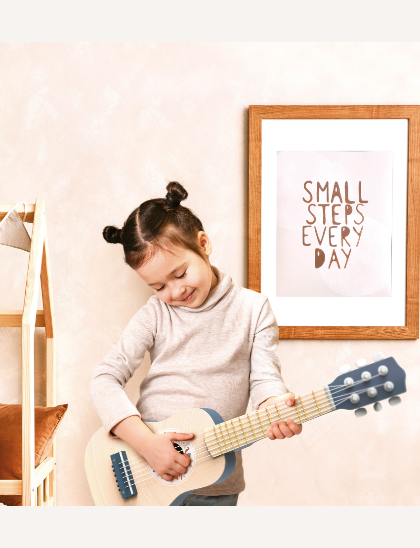 A young child with two small hair buns is playing a Speedy Monkey guitar. They are smiling and standing in a room with a framed sign on the wall that reads, "Small Steps Every Day." The room has a warm color scheme with a wooden piece of furniture.