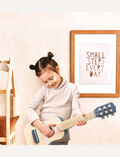 A young child with two small hair buns is playing a Speedy Monkey guitar. They are smiling and standing in a room with a framed sign on the wall that reads, 