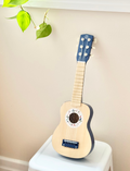 A young child with two small hair buns is playing a Speedy Monkey guitar. They are smiling and standing in a room with a framed sign on the wall that reads, 