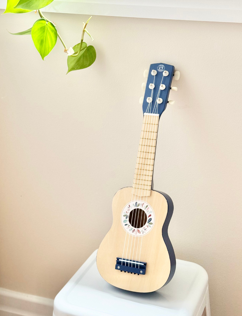 A young child with two small hair buns is playing a Speedy Monkey guitar. They are smiling and standing in a room with a framed sign on the wall that reads, "Small Steps Every Day." The room has a warm color scheme with a wooden piece of furniture.