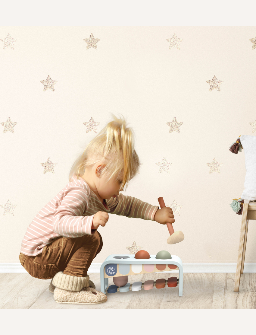 A child with blonde hair is squatting and playing with the Speedy Monkey Xylo Bench - Tap Tap Xylophone. The child is dressed in a striped long-sleeve shirt, brown pants, and beige slippers, set against a backdrop of a light-colored wall adorned with star patterns.