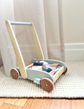 The Speedy Monkey Block Walker - 27 blocks is a charming wooden toy cart made from sustainably sourced wood, featuring four red wheels and filled with colorful geometric building blocks, such as triangles, squares, rectangles, and circles. The light blue handle complements the cart's sides, making it perfect for playtime adventures.