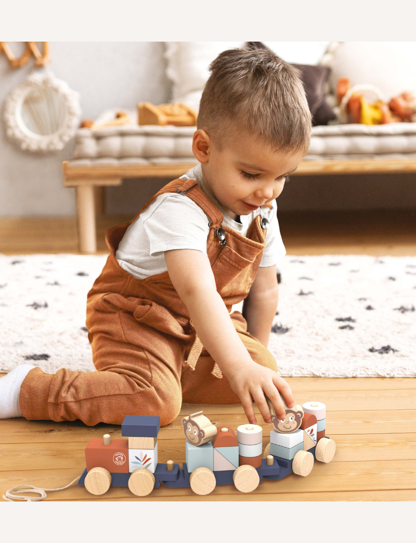 A vibrant Stacking Train by Speedy Monkey is spread across a white surface. The set features various blocks, including a piece with a monkey face, cylindrical shapes, and bright rectangular blocks. Some pieces have wheels attached and are crafted from sustainably sourced wood.