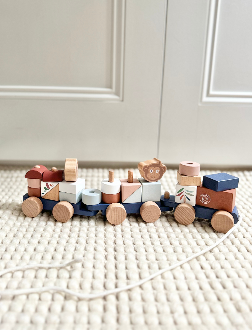 A vibrant Stacking Train by Speedy Monkey is spread across a white surface. The set features various blocks, including a piece with a monkey face, cylindrical shapes, and bright rectangular blocks. Some pieces have wheels attached and are crafted from sustainably sourced wood.