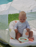 A baby sits in a shaded Sunnylife Baby Playmat with Shade in Apple Sorbet Multi on the grass, wearing a white onesie with small patterns. The baby holds a white bottle and a light blue ring, with a pool visible in the background.