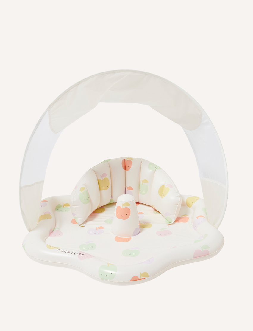 A baby sits in a shaded Sunnylife Baby Playmat with Shade in Apple Sorbet Multi on the grass, wearing a white onesie with small patterns. The baby holds a white bottle and a light blue ring, with a pool visible in the background.