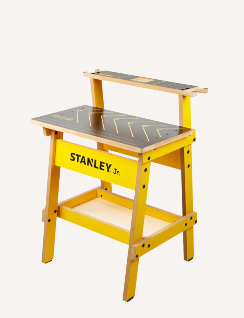 A child, equipped with safety goggles and a tool belt, is busy constructing a wooden model at a yellow Kids Workbench by Stanley, which has various tools like screwdrivers and clamps attached. The child is currently using a screwdriver to work on the model.