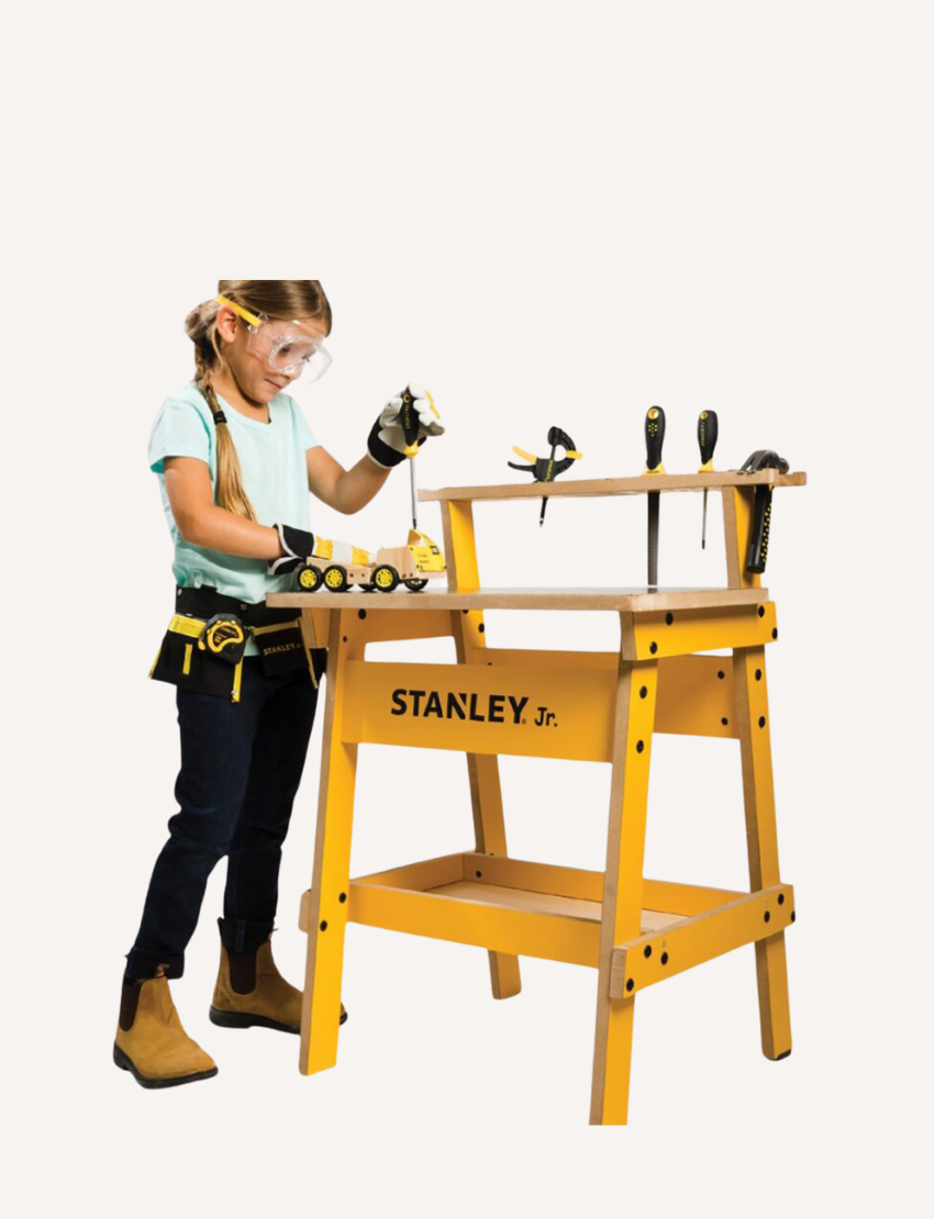 A child, equipped with safety goggles and a tool belt, is busy constructing a wooden model at a yellow Kids Workbench by Stanley, which has various tools like screwdrivers and clamps attached. The child is currently using a screwdriver to work on the model.