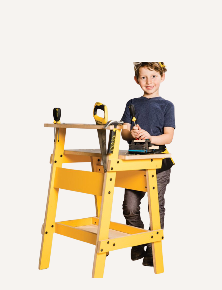 A child, equipped with safety goggles and a tool belt, is busy constructing a wooden model at a yellow Kids Workbench by Stanley, which has various tools like screwdrivers and clamps attached. The child is currently using a screwdriver to work on the model.