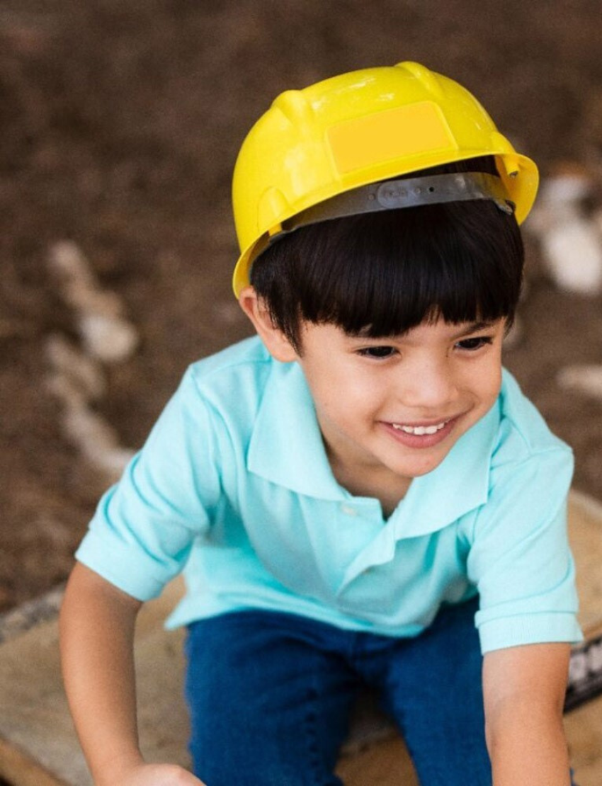 A child, dreaming of becoming a builder, wears a Stanley Construction Helmet and a light blue polo shirt, their face alight with happiness. The out-of-focus backdrop suggests an outdoor environment with dirt, sparking visions of upcoming construction adventures.