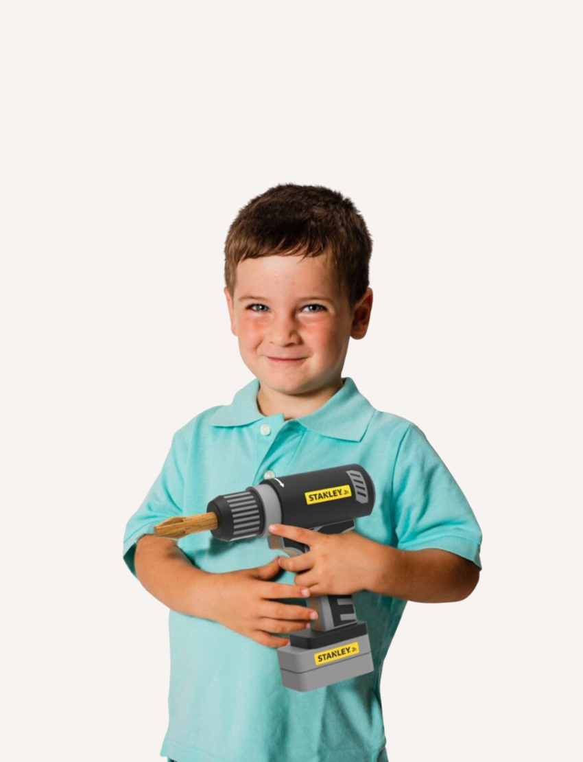 A cheerful child in a teal shirt holds a toy power drill from the "Wooden Tool Set - 28pcs" collection, with a wooden cork acting as the drill bit. The brand name "Stanley" is printed on the drill.