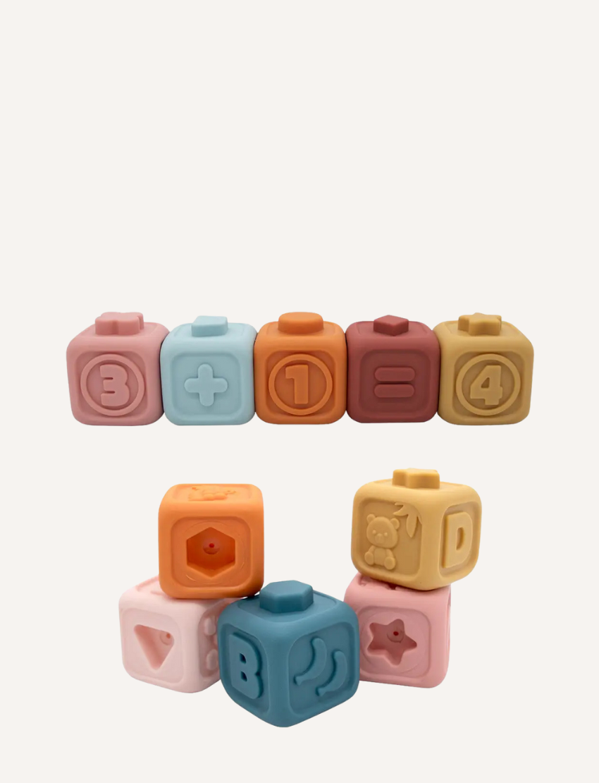 The image displays two collections of "My First Learning Blocks - Multi" by Playground. The top row contains six blocks in colors like pink, blue, orange, and brown featuring numbers and math symbols. The bottom row is a stack consisting of four blocks adorned with different shapes and letters.