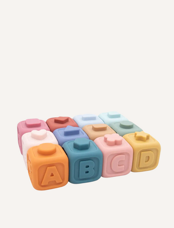 The image displays two collections of "My First Learning Blocks - Multi" by Playground. The top row contains six blocks in colors like pink, blue, orange, and brown featuring numbers and math symbols. The bottom row is a stack consisting of four blocks adorned with different shapes and letters.