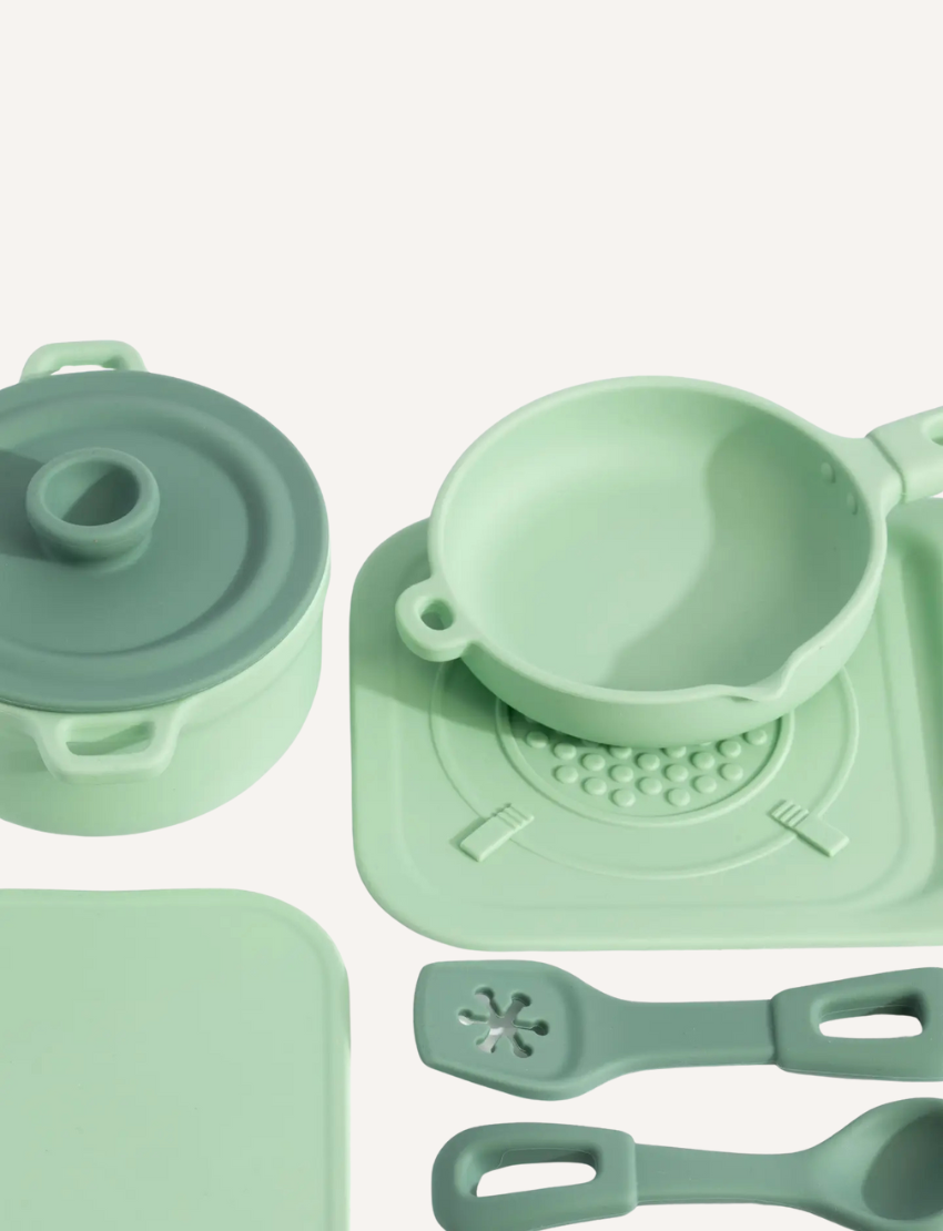 The Silicone Kitchen Playset by Playground features a selection of pastel green toy kitchen utensils, including a saucepan, a pot with a lid, a spatula, and a ladle—all arranged on a matching silicone mat. The items are showcased neatly against a simple backdrop.