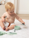 The Silicone Kitchen Playset by Playground features a selection of pastel green toy kitchen utensils, including a saucepan, a pot with a lid, a spatula, and a ladle—all arranged on a matching silicone mat. The items are showcased neatly against a simple backdrop.