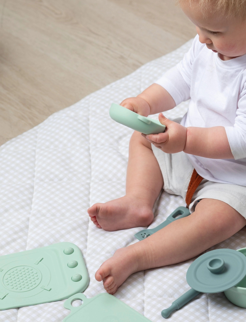 The Silicone Kitchen Playset by Playground features a selection of pastel green toy kitchen utensils, including a saucepan, a pot with a lid, a spatula, and a ladle—all arranged on a matching silicone mat. The items are showcased neatly against a simple backdrop.