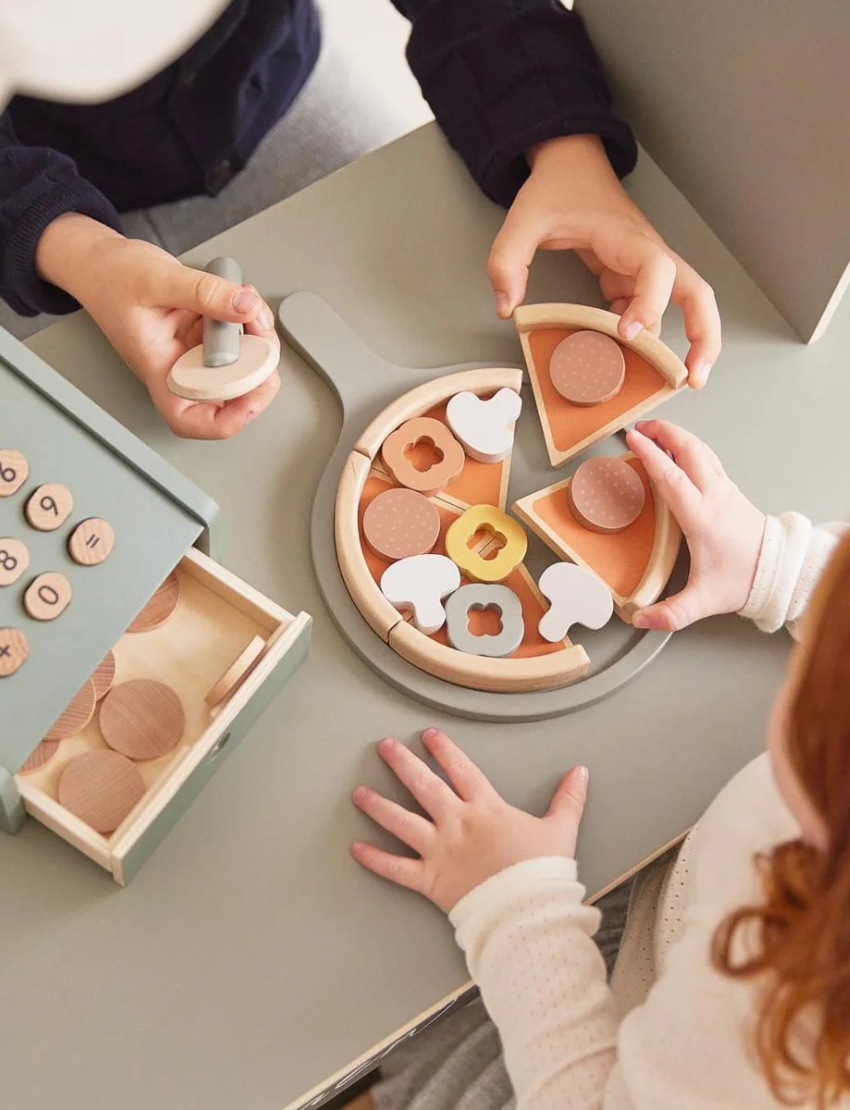 Children enjoying the Timber Pizza Set by Flexa. The set features a pan, pizza slices, and a variety of toppings. One child is using a spatula to hold a slice, while the other arranges toppings. A box containing extra pieces sits nearby on the table.