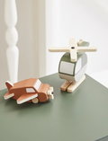 The Flexa Wooden Helicopter and Plane set features a green and white helicopter with wooden propeller and skids, alongside a brown airplane equipped with a wooden propeller and wheels. Both toys boast a minimalist design.