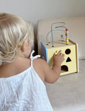 A blond toddler in a white sleeveless dress plays with a Moover Mini Activity Cube, featuring shaped holes and a wire maze with beads on top, set on a light-colored couch.