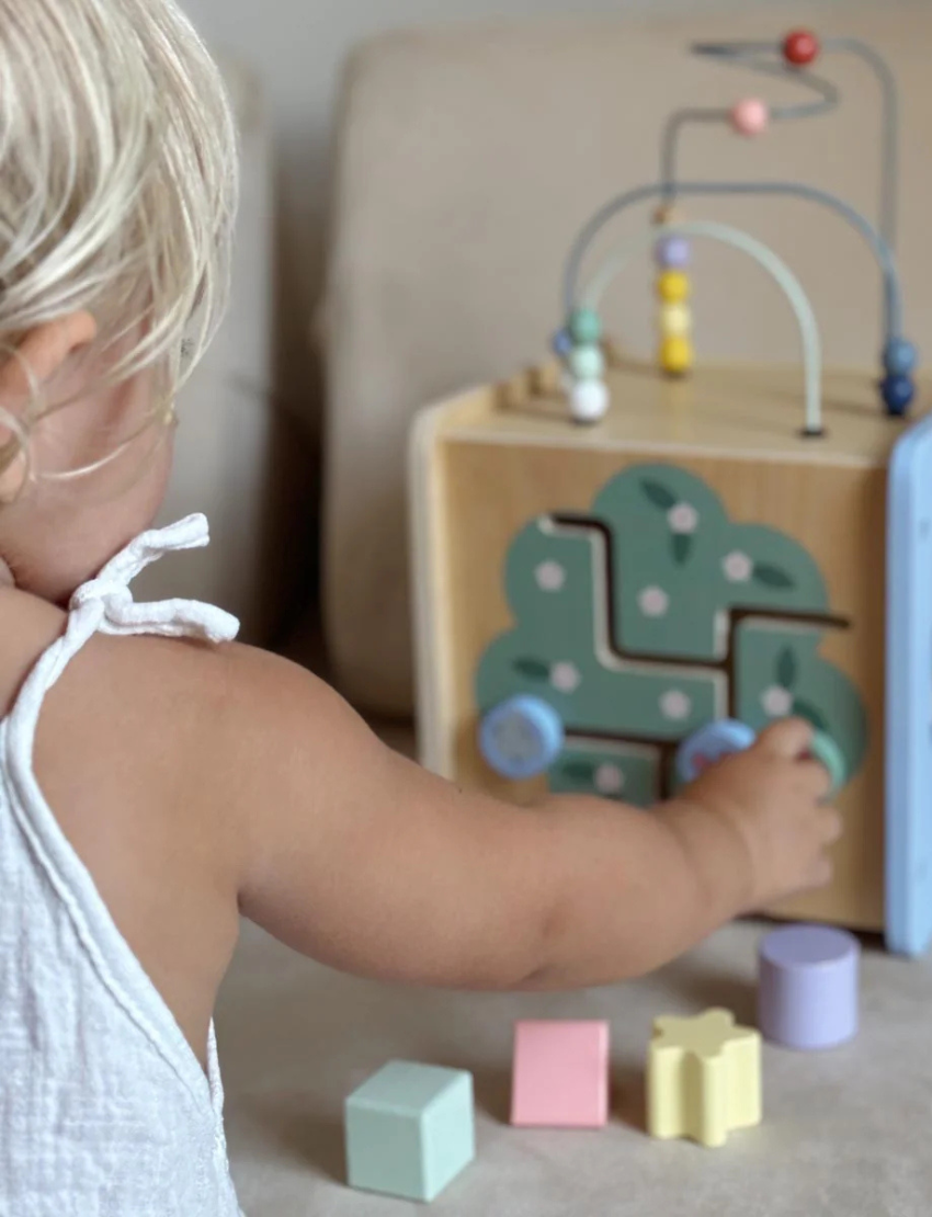 A blond toddler in a white sleeveless dress plays with a Moover Mini Activity Cube, featuring shaped holes and a wire maze with beads on top, set on a light-colored couch.