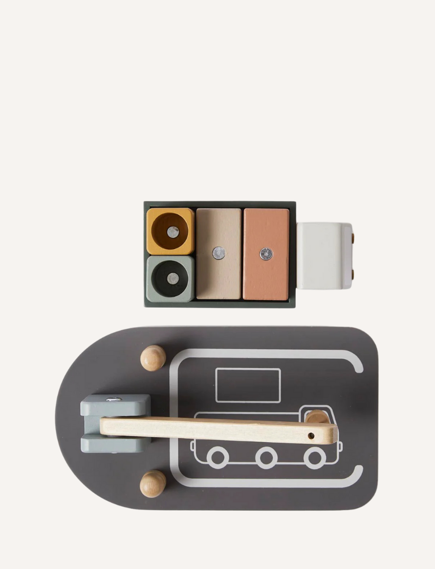 The Flexa Wooden Truck and Crane set includes a truck with block cargo, a balance scale, and two bins marked with recycling symbols. The set features a gray base designed with road patterns and comes with a wooden lever arm. It showcases muted tones of green, brown, and gray.