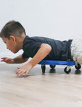 The Play Way's Regulation Through Movement Kit is opened, showcasing a hammock swing with a blue edge, a gray exercise ball, and a yellow scooter board with four casters. Various accessories for sensory regulation can be seen partly inside and around the green and blue box.