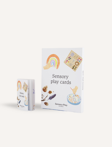 Two sets of "Sensory Play Card Set" by Sensory Play Australia are displayed, inviting sensory exploration and emotional regulation. The larger set stands upright, featuring illustrations of a rainbow, wooden blocks, painted stones, and a sensory rice bowl. The smaller set is stacked neatly with similar designs visible on the box.