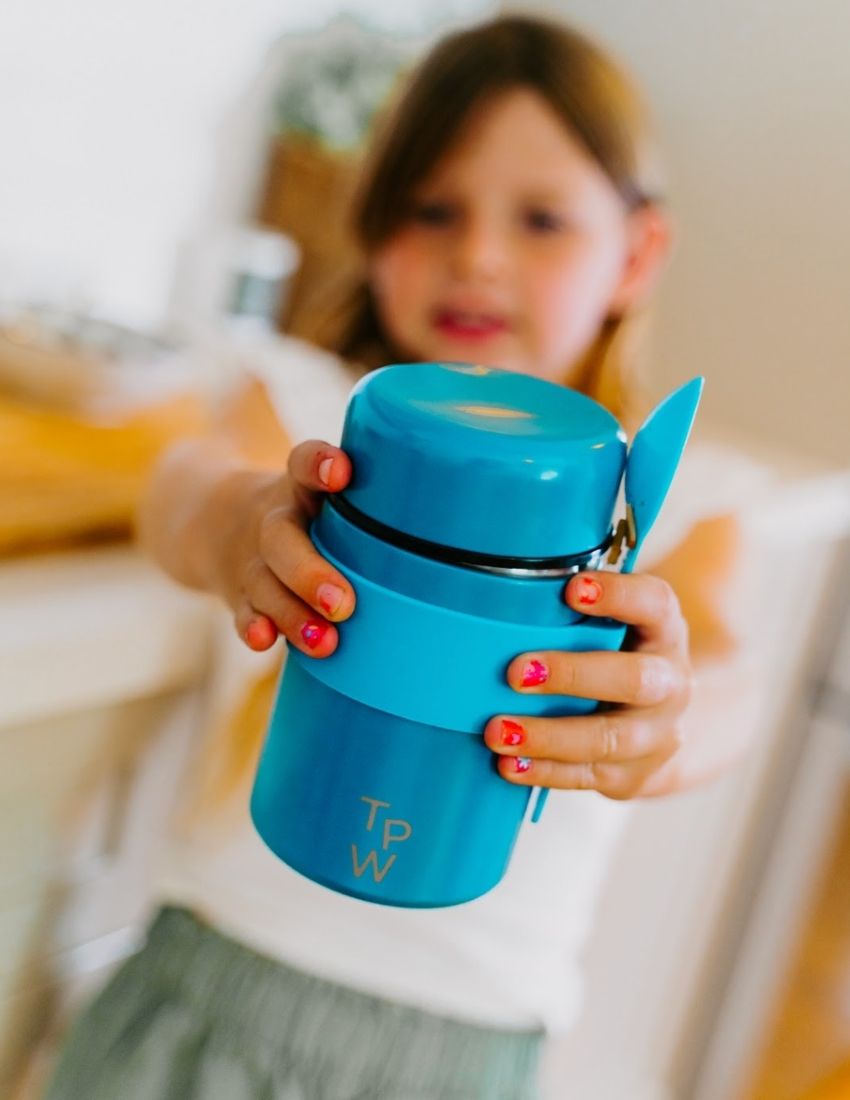 A blue insulated food jar from The Play Way with the initials "T P W" printed on the front. The Lunch Buddy Insulated Container Set features a matching blue silicone band around its midsection that holds a blue spoon, along with a leak-proof lid. The background is plain white.