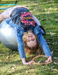 The Play Way's Regulation Through Movement Kit is opened, showcasing a hammock swing with a blue edge, a gray exercise ball, and a yellow scooter board with four casters. Various accessories for sensory regulation can be seen partly inside and around the green and blue box.