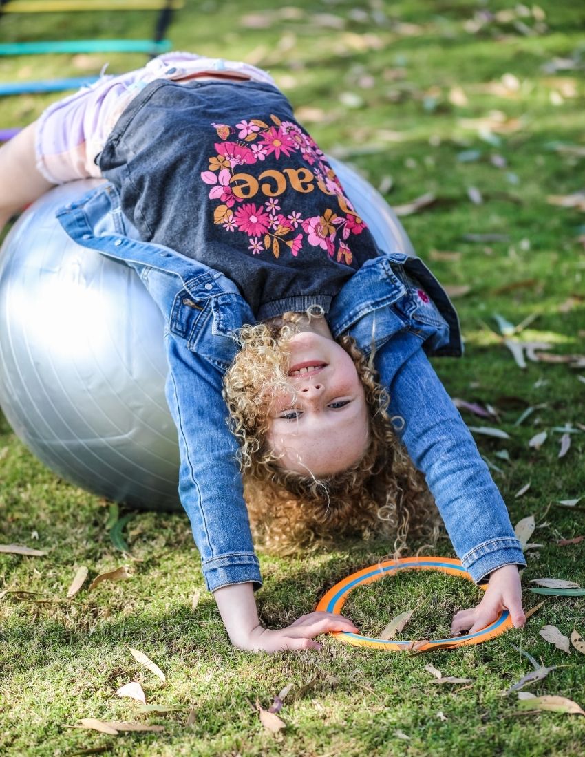 The Play Way's Regulation Through Movement Kit is opened, showcasing a hammock swing with a blue edge, a gray exercise ball, and a yellow scooter board with four casters. Various accessories for sensory regulation can be seen partly inside and around the green and blue box.