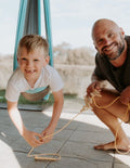 The Play Way's Regulation Through Movement Kit is opened, showcasing a hammock swing with a blue edge, a gray exercise ball, and a yellow scooter board with four casters. Various accessories for sensory regulation can be seen partly inside and around the green and blue box.