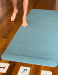 A rolled-up large turquoise yoga mat from The Play Way brand, secured with black straps. The text 