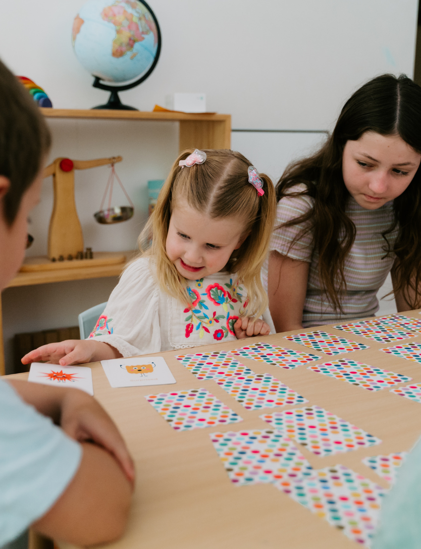 The "Emotions Buddy Naming Feelings Set" by Sensory Play Australia is a colorful box set for children, featuring an empathy-building emotions check-in chart, playing cards labeled "Emotions," a guidebook, and illustrated emotion-themed materials. The packaging is decorated with vibrant rainbow designs.