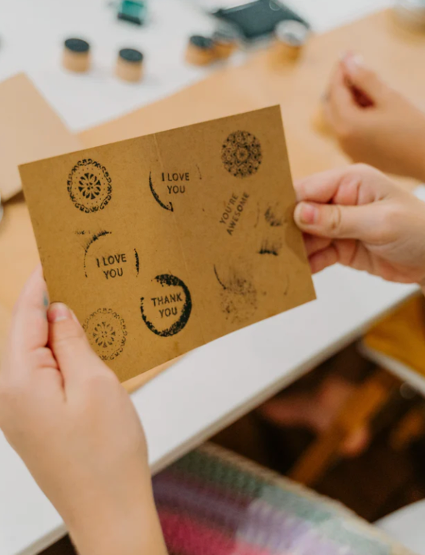 A flat lay display featuring the Poppy & Daisy DIY Gifting Set on a white surface showcases an enchanting collection for a delightful crafting experience. This set includes four rubber stamps with various messages, a vibrant green ink pad, a roll of brown paper, handmade cards with matching envelopes, a spool of twine, and small branches adorned with delicate white flowers.