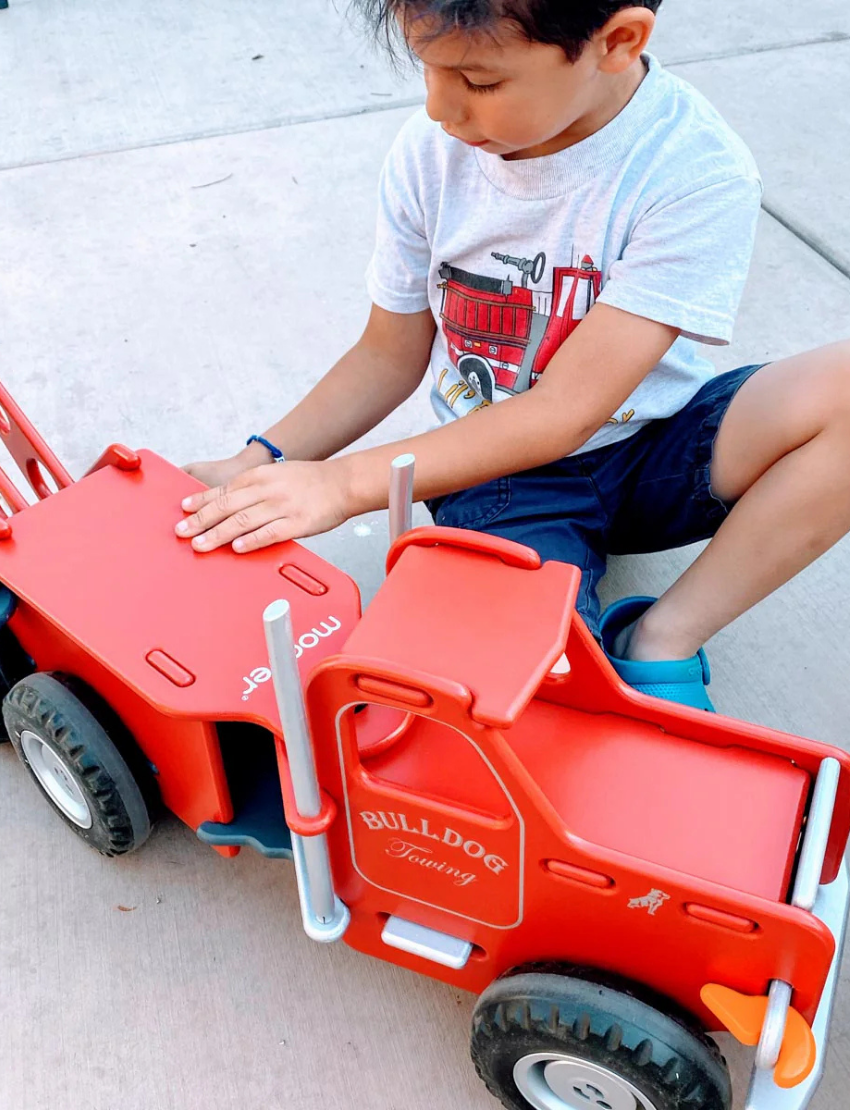 The Moover Mack Ride On Truck in red boasts six black wheels, a rear hook, and features "Mack" on the hood with "Bulldog Towing" on the side. This durable miniature model stands out against a white backdrop, highlighting its robust design.