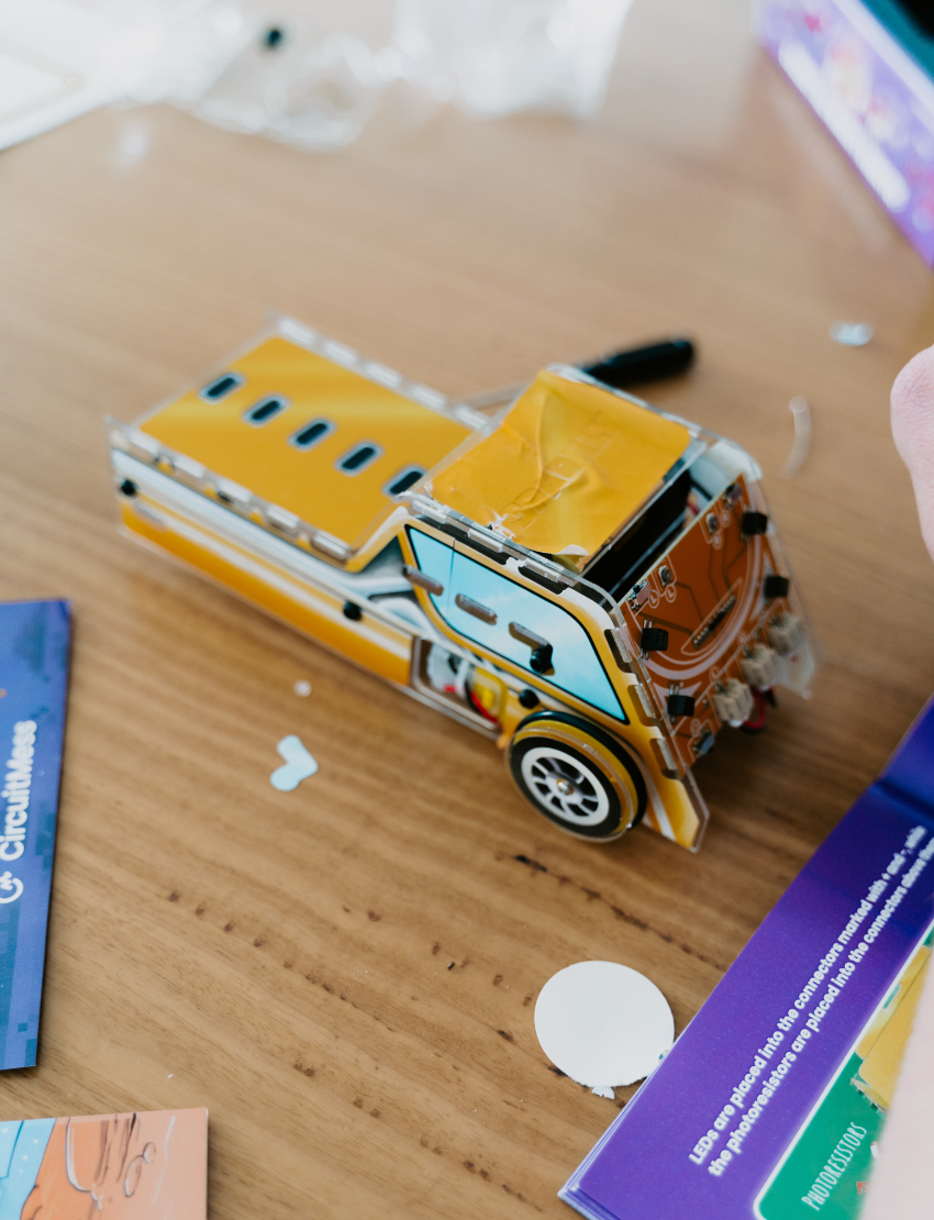 A toy robot car kit called "STEM Adventure - Sparkly Robot Car" by CircuitMess is displayed. The assembled car, in yellow and blue, sits in front of its packaging box, which features a car illustration and notes it is suitable for ages 7 and up.