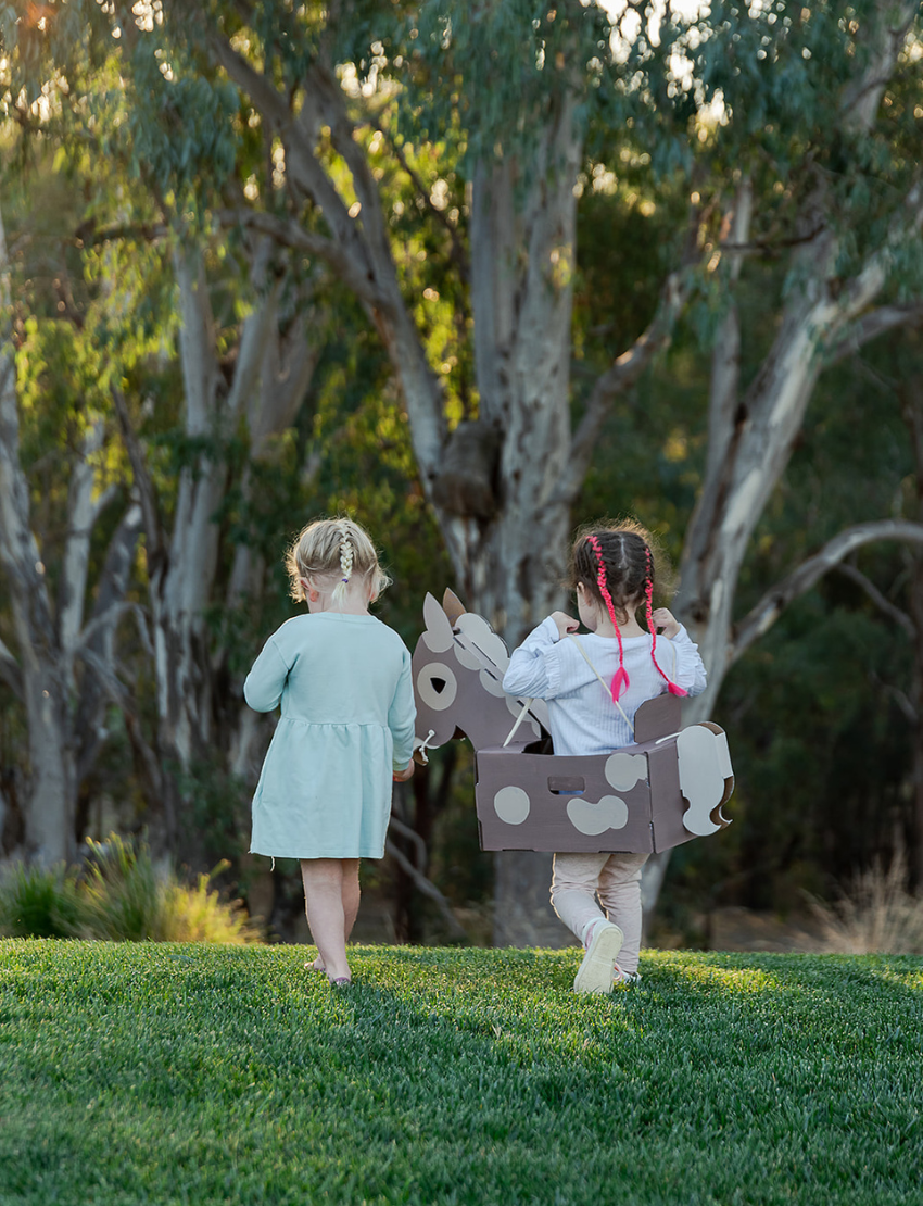 Two young children are playing with a cardboard fort. One child stands inside holding a toy sword, while the other stands beside the fort, raising a toy sword and holding a Horse Costume by Les Petite Artistes. The fort is decorated to resemble a medieval castle, setting the stage for their artistic adventures.