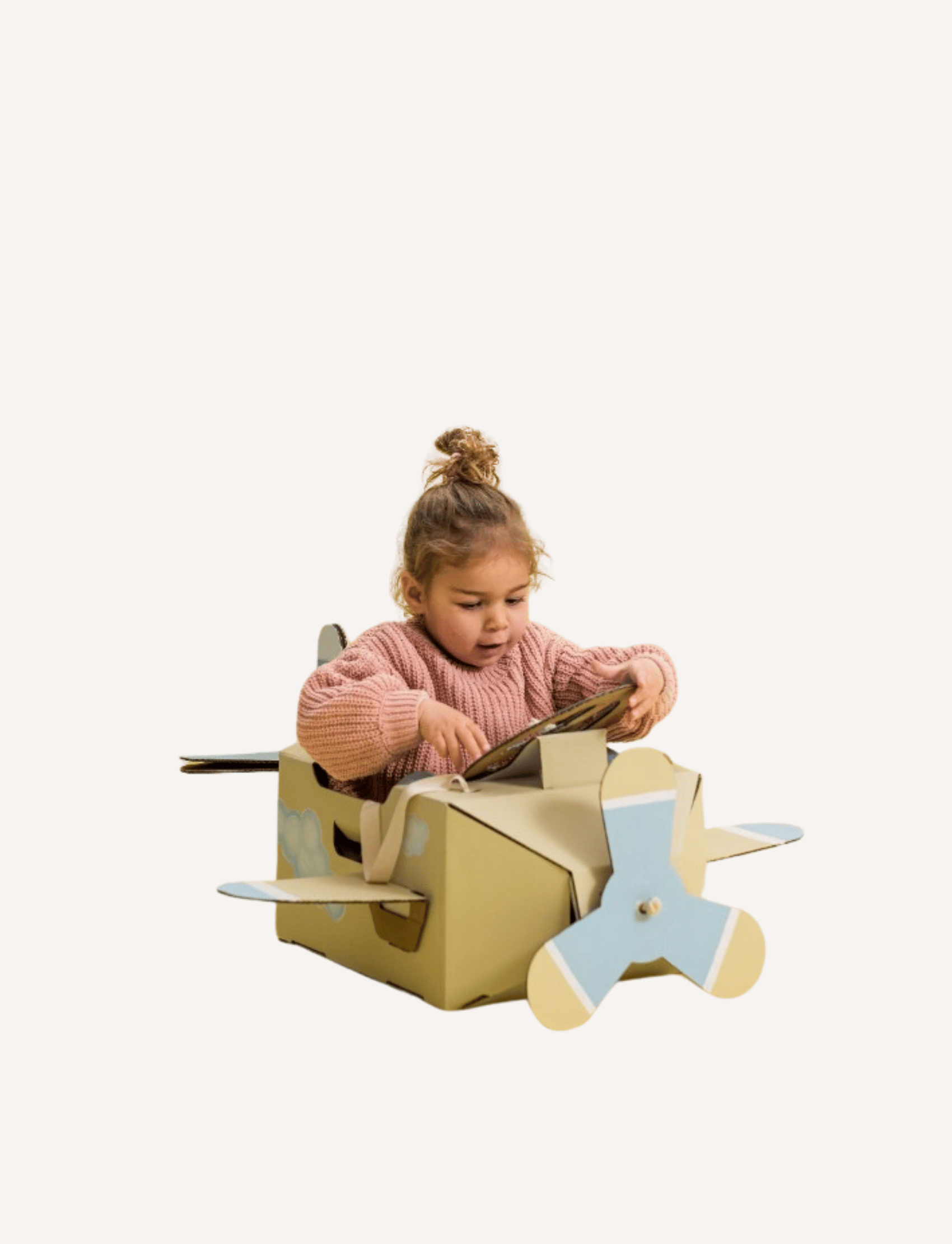 A young child with light brown hair in a bun, wearing a pink sweater and blue jeans, stands barefoot and looks down at a Les Petite Artistes Jet Plane Costume shaped like an airplane with blue propellers on a plain, white background. It's clear that young aviators can foster their artistic dreams from an early age.