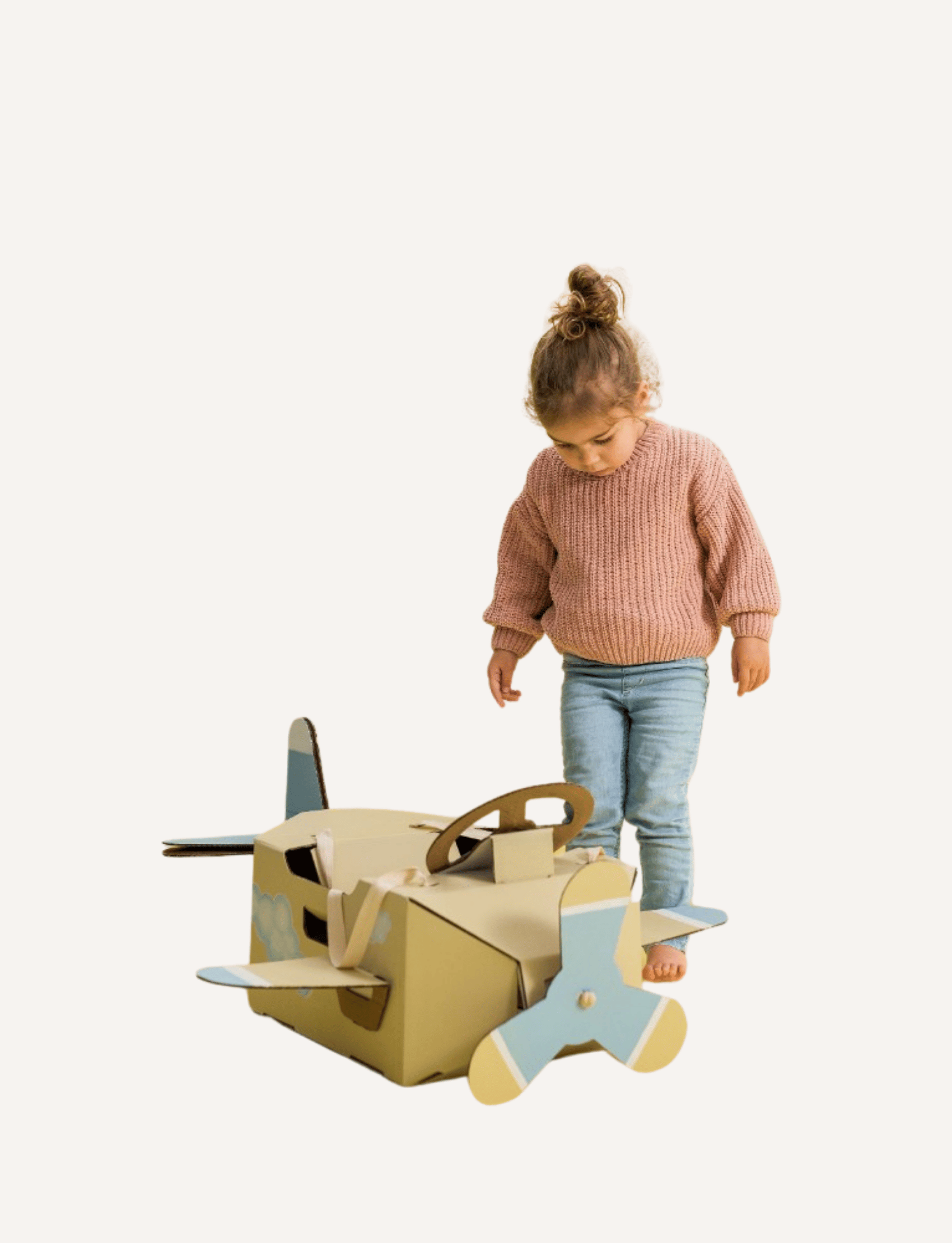 A young child with light brown hair in a bun, wearing a pink sweater and blue jeans, stands barefoot and looks down at a Les Petite Artistes Jet Plane Costume shaped like an airplane with blue propellers on a plain, white background. It's clear that young aviators can foster their artistic dreams from an early age.