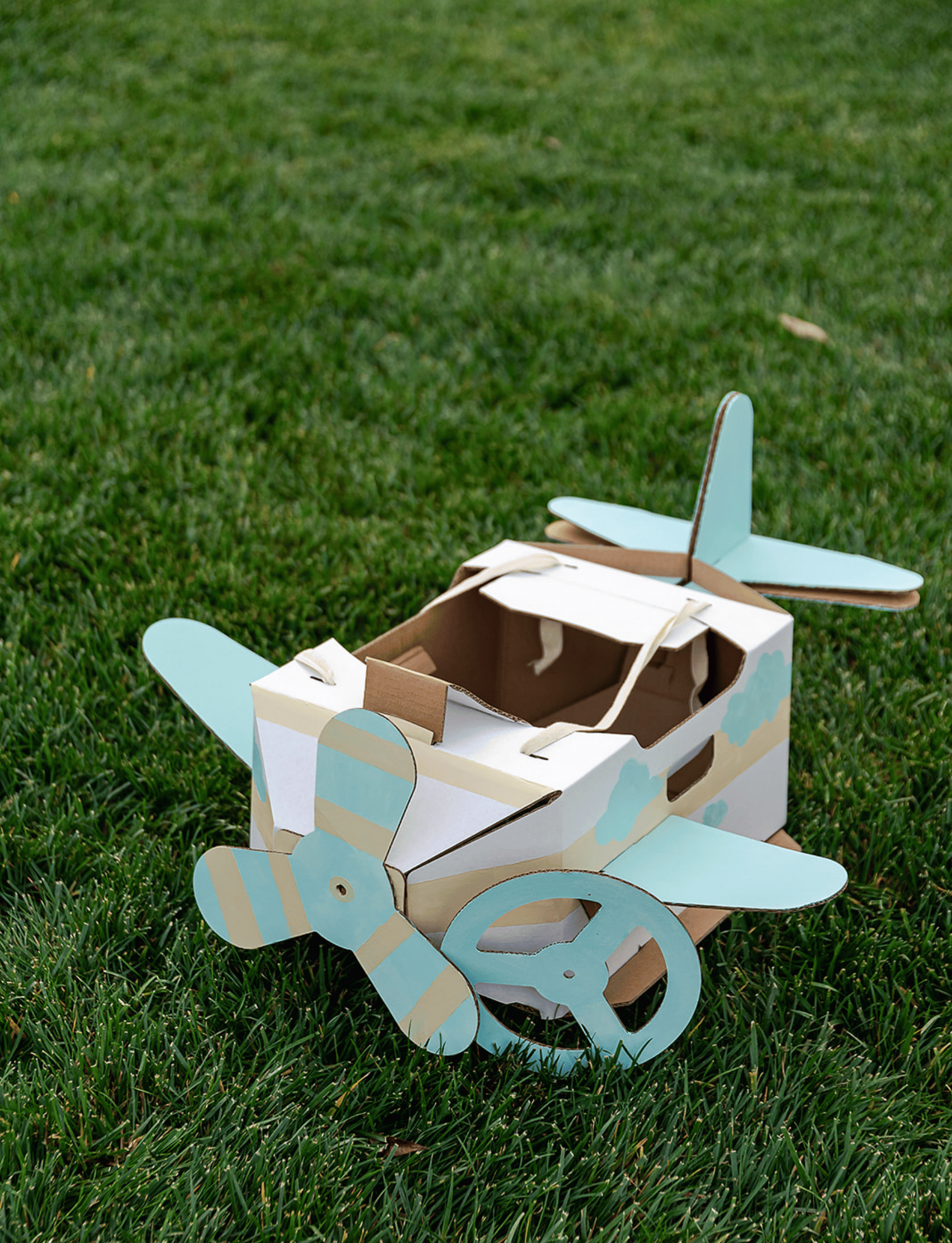 A young child with light brown hair in a bun, wearing a pink sweater and blue jeans, stands barefoot and looks down at a Les Petite Artistes Jet Plane Costume shaped like an airplane with blue propellers on a plain, white background. It's clear that young aviators can foster their artistic dreams from an early age.