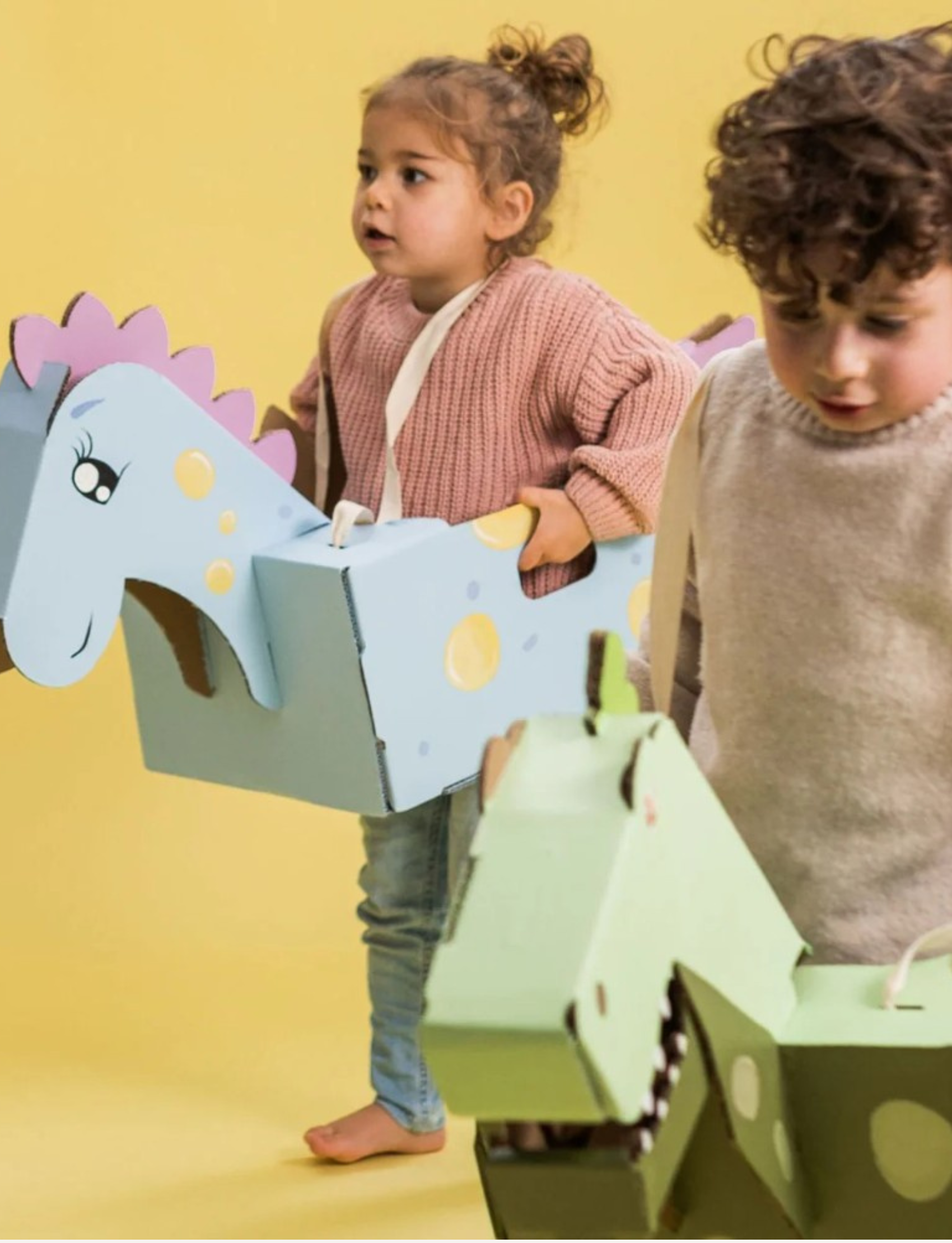 Two young children embark on prehistoric adventures with wearable cardboard animal costumes from Les Petite Artistes. The child on the left, in a pink sweater and blue jeans, holds a blue costume resembling a horse. The child on the right, wearing a beige sweater, joyfully brandishes their Nice Dinosaur Costume.